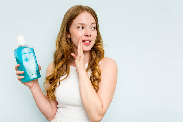 Молодая белая женщина, держащая в руках жидкость для полоскания рта, изолированную на синем фоне, говорит тайные горячие новости тормозов и смотрит в сторону - Фото, изображение