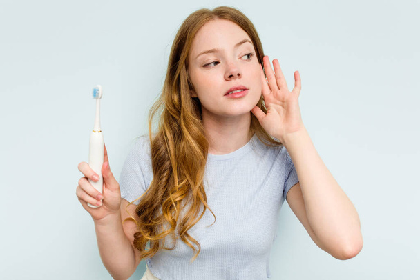 青の背景に孤立した電動歯ブラシを持っている若い白人女性はゴシップを聞いてみる. - 写真・画像