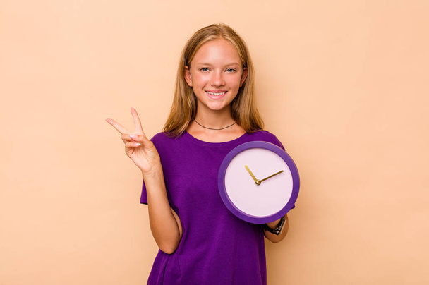 Niña caucásica sosteniendo un reloj aislado sobre fondo beige alegre y despreocupada mostrando un símbolo de paz con los dedos. - Foto, Imagen