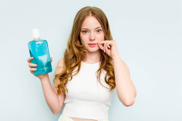 Молода біла жінка тримає полоскання рота ізольовано на синьому фоні з пальцями на губах тримаючи секрет
. - Фото, зображення
