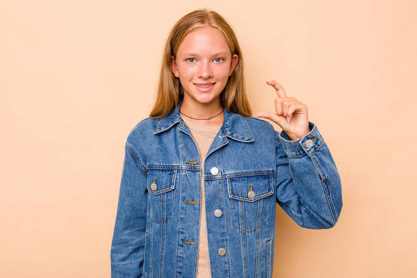 Caucasian teen girl isolated on beige background holding something little with forefingers, smiling and confident. - Photo, Image