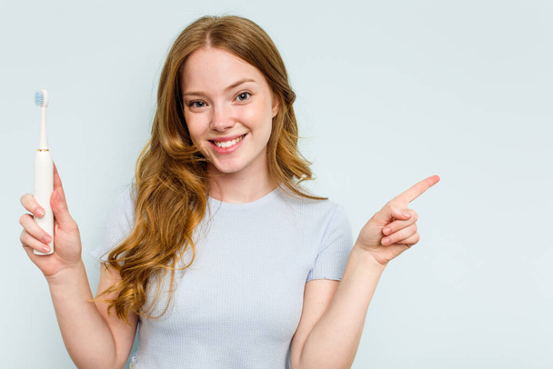 Young caucasian woman holding electric toothbrush isolated on blue background smiling and pointing aside, showing something at blank space. - Photo, image