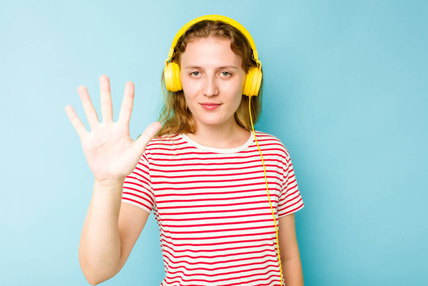 Young caucasian woman wearing headphones isolated on blue background smiling cheerful showing number five with fingers. - Photo, Image