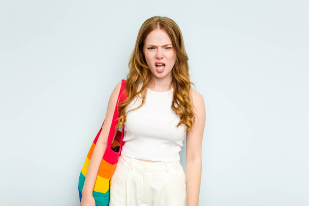 jonge blanke vrouw houden LGTBI tas geïsoleerd op blauwe achtergrond schreeuwen zeer boos en agressief. - Foto, afbeelding