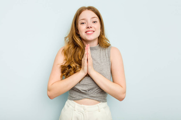 口の近くで祈る手を握って青い背景に隔離された若い白人女性は、自信を感じている. - 写真・画像