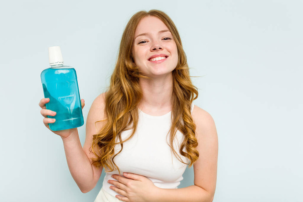 Młoda kaukaska kobieta trzyma płyn do płukania ust odizolowany na niebieskim tle śmiejąc się i bawiąc. - Zdjęcie, obraz