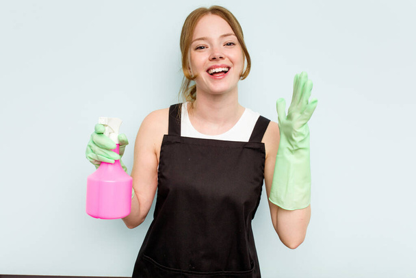 Νεαρή καθαρίστρια, απομονωμένη σε μπλε φόντο, δέχεται μια ευχάριστη έκπληξη, ενθουσιασμένη και σηκώνει τα χέρια.. - Φωτογραφία, εικόνα