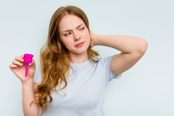 Junge kaukasische Frau hält Menstruationstasse isoliert auf blauem Hintergrund und berührt Hinterkopf, denkt nach und trifft eine Wahl. - Foto, Bild