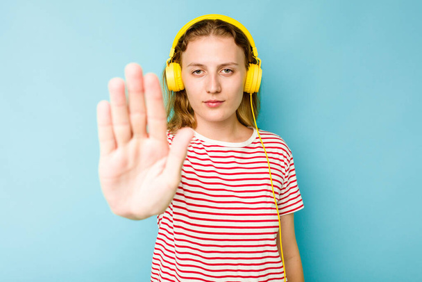 Young caucasian woman wearing headphones isolated on blue background standing with outstretched hand showing stop sign, preventing you. - Photo, Image