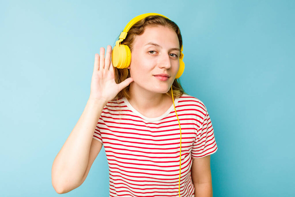 Nuori valkoihoinen nainen yllään kuulokkeet eristetty sinisellä taustalla yrittää kuunnella juoruja. - Valokuva, kuva