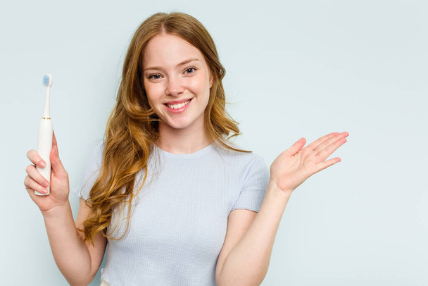 青い背景に孤立した電動歯ブラシを持っている若い白人女性は、手のひらにコピースペースを示し、腰に別の手を保持します. - 写真・画像