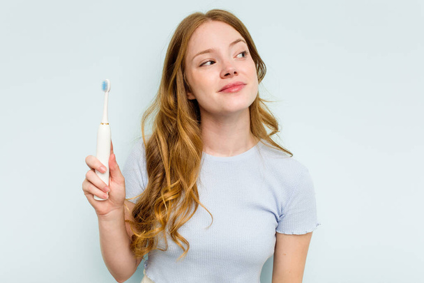 Mavi arka planda elektrikli diş fırçası tutan beyaz bir kadın hedeflerine ve amaçlarına ulaşma hayali kuruyor. - Fotoğraf, Görsel