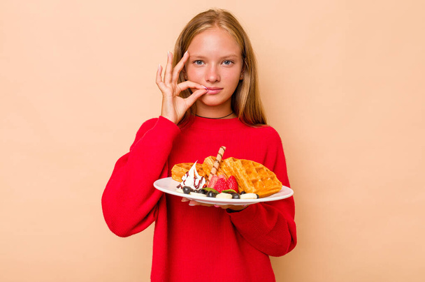 Little caucasian girl holding a waffles isolated on beige background with fingers on lips keeping a secret. - Photo, Image