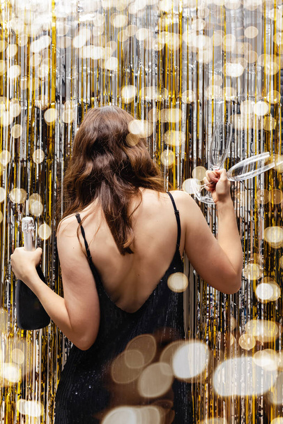 Woman entering new year party with bottle of champagne and glasses in her hands. Festive shiny curtains and blurred lights - Foto, Imagem