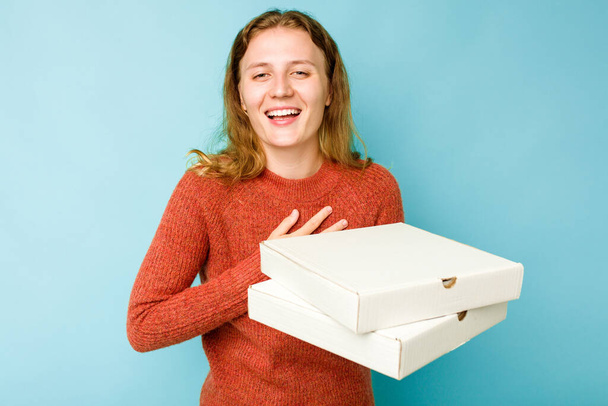 Νεαρή λευκή γυναίκα που κρατάει κουτιά πίτσας απομονωμένα σε μπλε φόντο γελάει δυνατά κρατώντας το χέρι στο στήθος. - Φωτογραφία, εικόνα