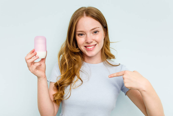 Mujer caucásica joven sosteniendo desodorante aislado sobre fondo azul persona señalando a mano a un espacio de copia de la camisa, orgulloso y seguro - Foto, imagen