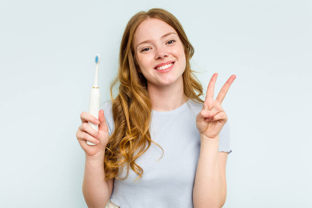 Junge kaukasische Frau hält elektrische Zahnbürste isoliert auf blauem Hintergrund und zeigt fröhlich und unbeschwert ein Friedenssymbol mit Fingern. - Foto, Bild