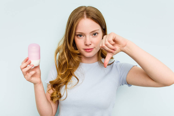 Young caucasian woman holding deodorant isolated on blue background showing a dislike gesture, thumbs down. Disagreement concept. - Foto, Imagen