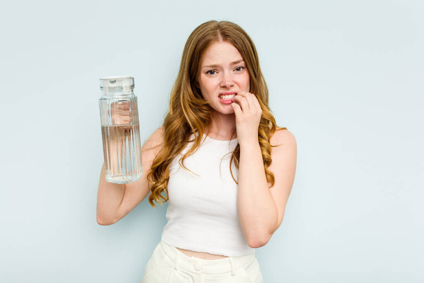 Giovane donna caucasica in possesso di vaso d'acqua isolato su sfondo blu mordere le unghie, nervoso e molto ansioso. - Foto, immagini