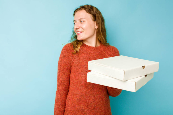 Young caucasian woman holding pizzas boxes isolated on blue background looks aside smiling, cheerful and pleasant. - Фото, изображение