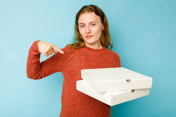 Νεαρή Καυκάσια γυναίκα που κρατάει κουτιά πίτσας απομονωμένα σε μπλε φόντο άτομο που δείχνει με το χέρι σε χώρο φωτοαντιγραφής πουκάμισου, περήφανη και με αυτοπεποίθηση - Φωτογραφία, εικόνα