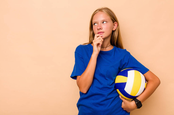 Little caucasian girl playing volleyball isolated on beige background looking sideways with doubtful and skeptical expression. - 写真・画像