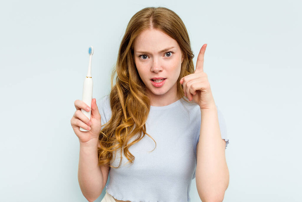 Молодая белая женщина с электрической зубной щеткой на голубом фоне, имеющая идею, концепцию вдохновения. - Фото, изображение