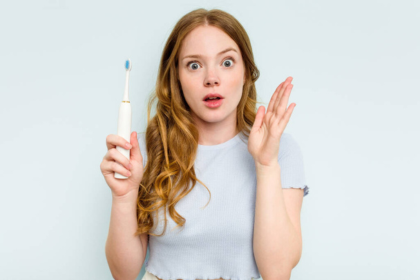 青の背景に隔離された電動歯ブラシを持っている若い白人女性は驚いてショックを受けました. - 写真・画像