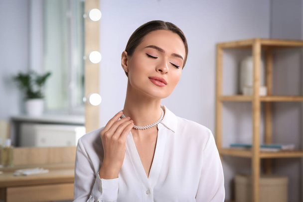 Young woman trying on elegant pearl necklace indoors - Фото, изображение