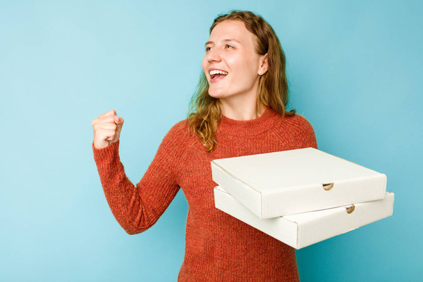 Nuori valkoihoinen nainen tilalla pizzat laatikot eristetty sininen tausta nostaa nyrkki voiton jälkeen, voittaja käsite. - Valokuva, kuva