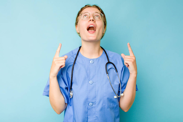 Nuori sairaanhoitaja valkoihoinen nainen eristetty sinisellä taustalla osoittaa ylösalaisin suu auki. - Valokuva, kuva
