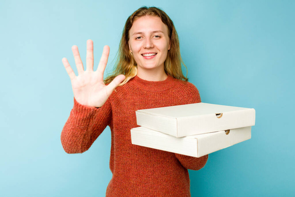 Νεαρή Καυκάσια γυναίκα που κρατάει κουτιά πίτσας απομονωμένα σε μπλε φόντο χαμογελώντας χαρούμενα δείχνοντας τον αριθμό πέντε με τα δάχτυλα. - Φωτογραφία, εικόνα