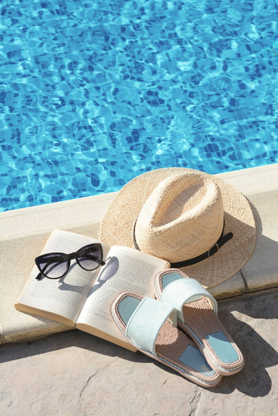 Stylish sunglasses, slippers, straw hat and book at poolside on sunny day. Beach accessories - Foto, Imagem