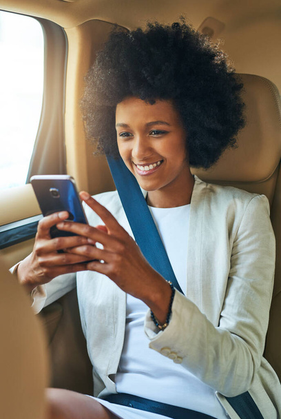 Ik vraag me af of ik moet reageren. een vrolijke jonge zakenvrouw bladert op haar mobiele telefoon terwijl ze zit in de passagiersstoel van een auto op weg naar haar werk - Foto, afbeelding