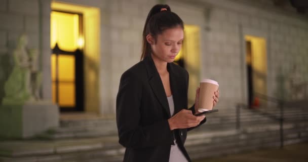 Vuosituhannen liiketoiminnan nainen juo kahvia ja käyttää älypuhelinta toimistonsa ulkopuolella yöllä. Omistautunut nuori ammattilainen lähettää tekstiviestin matkapuhelimeen. 4 k. - Materiaali, video