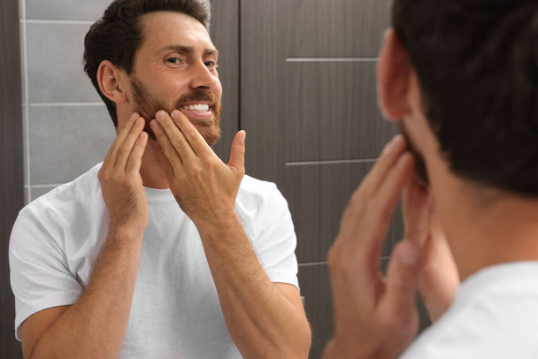 Smiling bearded man looking at mirror in bathroom - Фото, изображение