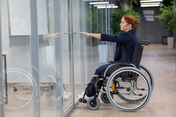 Рыжая белая женщина в инвалидном кресле пытается открыть дверь в офисе - Фото, изображение