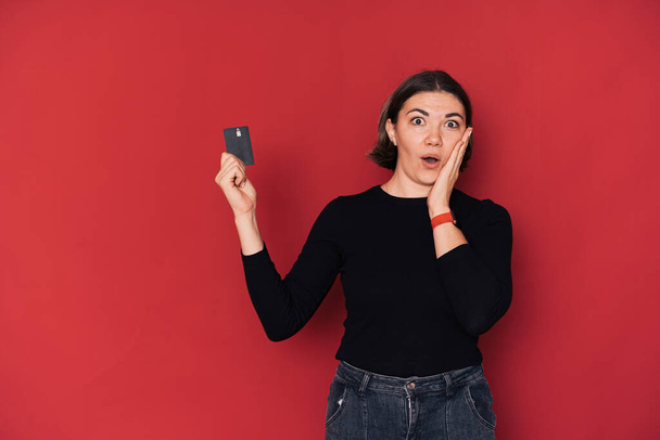 Lady in shock houden credit card in haar hand tegen rode achtergrond. - Foto, afbeelding