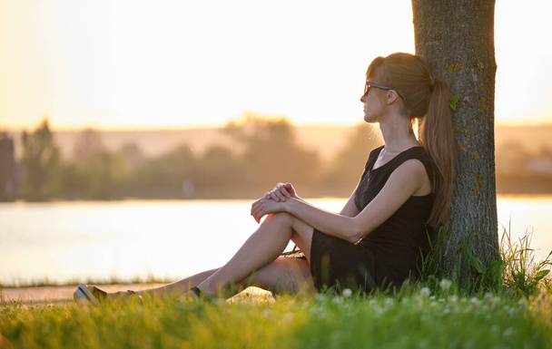 Eenzame jonge vrouw zit alleen op gras gazon aan de oever van het meer genieten van warme avond. Welzijn en ontspanning in de natuur concept. - Foto, afbeelding