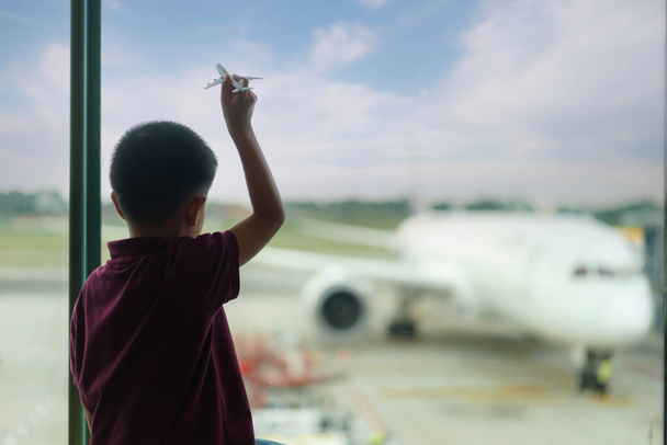 Asian Boy with airplane model at empty airport terminal waiting for departure looking out the window. Child in T-shirt and shorts stands at lounge waiting for plane flight. Family trip concept - Photo, Image