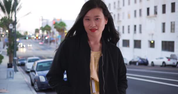 Mulher chinesa jovem em jaqueta de bombardeiro preto em pé na rua da cidade com as mãos nos bolsos. Asian millennial fica na calçada com cabelos longos soprando no vento. 4k - Filmagem, Vídeo
