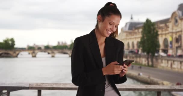 Mujer de negocios sonriente en el puente de París sobre el río Sena mensajes de texto con teléfono celular. Milenial profesional a la espera de paseo en la calle París envía un mensaje de texto al conductor. 4k - Imágenes, Vídeo