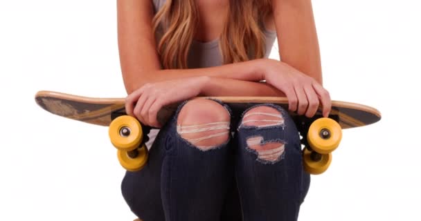 Gros plan de jeune femme blanche portant un jean déchiré et un débardeur assis, tenant skateboard sur fond blanc avec copyspace. Gros plan d'une jeune fille millénaire assise les bras croisés en studio. 4k - Séquence, vidéo