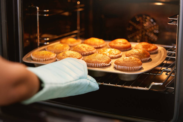 Hand, Ofen und Muffins backen, essen oder süße leckere Kuchen auf einem Blech zu Hause in der Küche zubereiten. Hände des Bäckers nehmen heißen Muffin gebackene Mahlzeit, Leckereien oder Freude an Gebäck machen im Haus. - Foto, Bild
