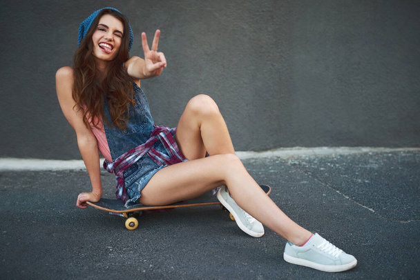 Taking her board out for a skate. Portrait of a cheerful young woman seated on a skateboard while showing the peace sign outside during the day - Foto, Imagen