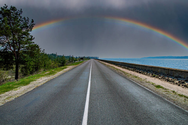 Un arco iris con una nube de trueno en el horizonte de una carretera. Arco iris en la tormenta. Asfalto pavimento. Marcas de carretera en asfalto. Horizonte nuboso. Camino costero. Vete de vacaciones. Temporada de verano. - Foto, Imagen