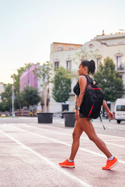 vertical photo of a latina sportswoman walking with a backpack on a city athletics track, urban sport and healthy lifestyle concept, copy space for text - Photo, Image