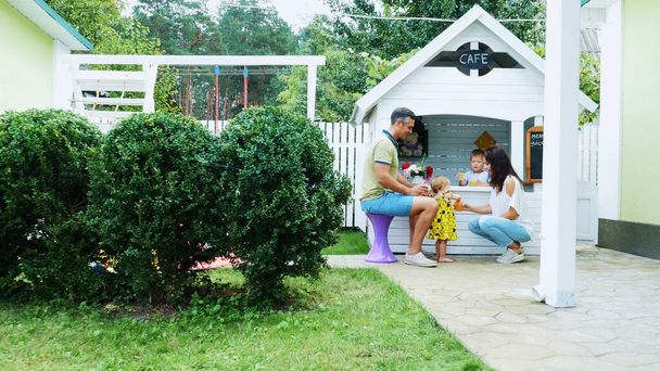 summer, in the garden, parents play with young children, a girl and a boy, in a cafe, in a childrens play house, treat children with freshly squeezed fruit juices, drink juices. High quality photo - Φωτογραφία, εικόνα