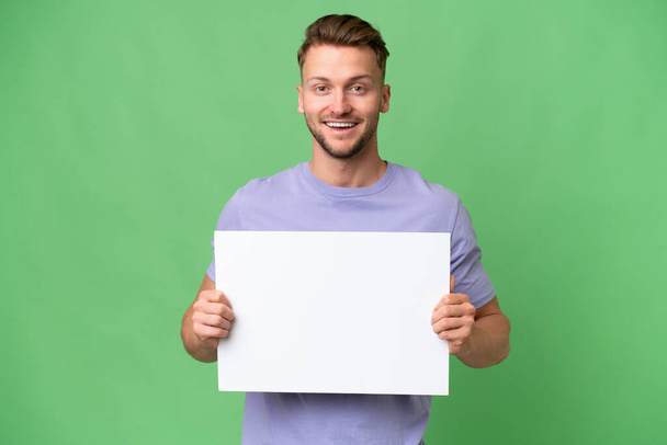 Νεαρός ξανθός καυκάσιος άντρας πάνω από απομονωμένο φόντο κρατώντας ένα άδειο πλακάτ με χαρούμενη έκφραση - Φωτογραφία, εικόνα