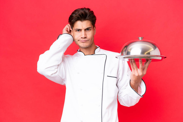 Mladý kavkazský kuchař muž drží zásobník izolované na červeném pozadí s pochybnostmi - Fotografie, Obrázek